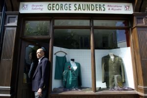 Older man in a tailored suit outside a tailor shop titled 'George Saunders'